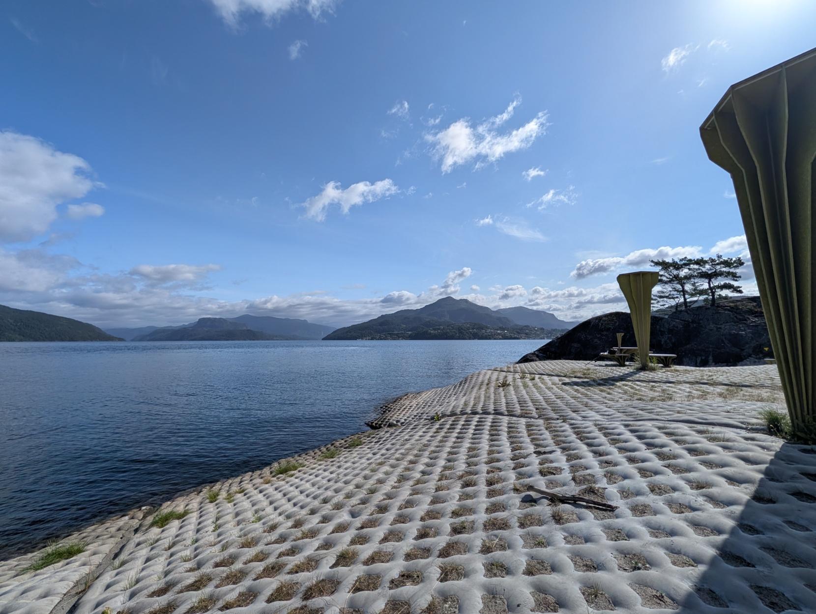 Frå Ropeid rasteplass: Eit breidvinkla bilde over fjorden under ein klår, blå himmel med lette skyer. I framgrunnen er bakken dekka av særmerkte, ruteforma betongmadrasser der graset gror i holromma. Til høgre står dei karakteristiske, lysegrøne sopp- eller vifteforma leskura i betong, i tillegg til sitjeplassar på svaberget. Til venstre opnar det mørkeblå vatnet seg ut mot fjella i bakgrunnen.
