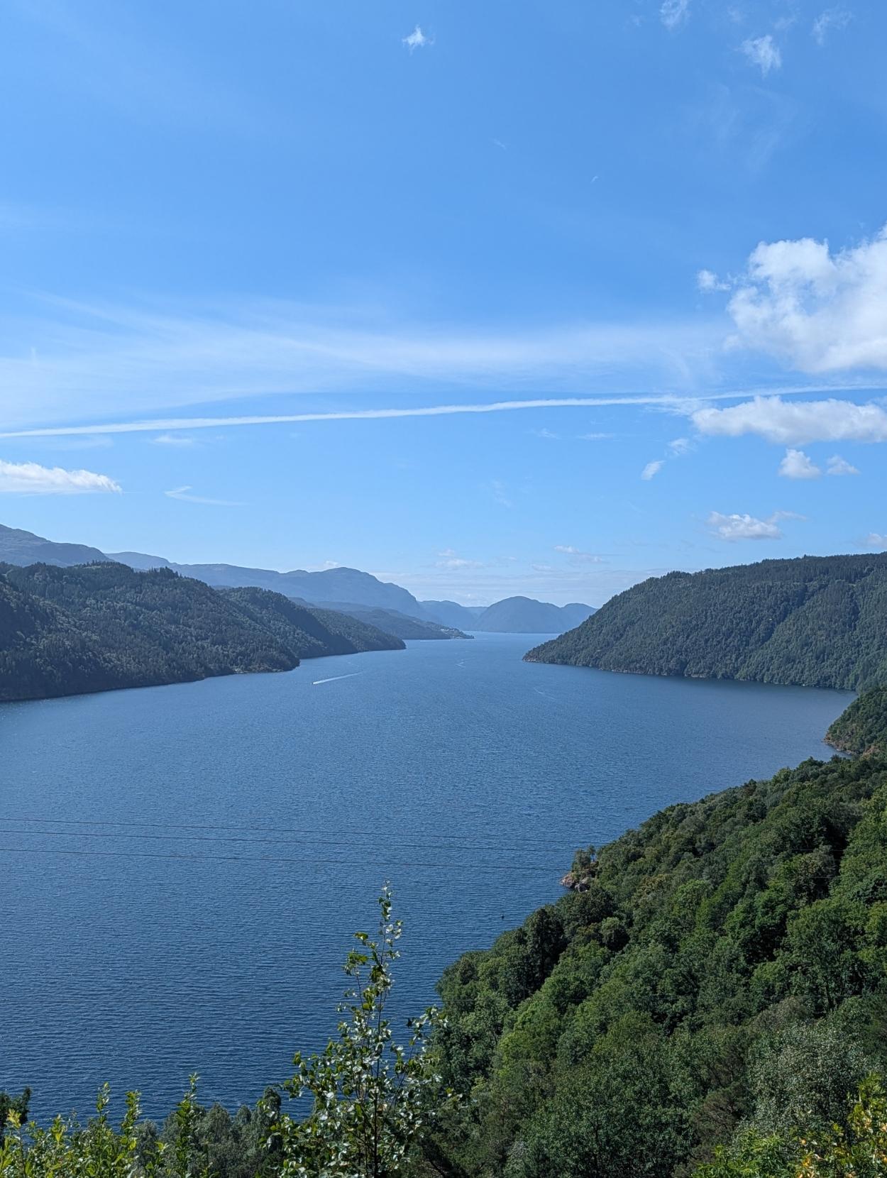 Utsikt sørover mot Sand frå Suldal: Eit høgt vertikalt panorama over ein brei, mørkeblå fjord som strekkjer seg innover i landskapet. Fjorden er omkransa av bratte, skogskledde åsar på begge sider, med tette grønne tretoppar i framgrunnen nede til høgre. I det fjerne aukar avstanden til blåaktige fjellkonturar. Over landskapet er det ein klår, solrik blå himmel med lette slørskyer og ein tynn, kvit flystripe som går vassrett over biletet.