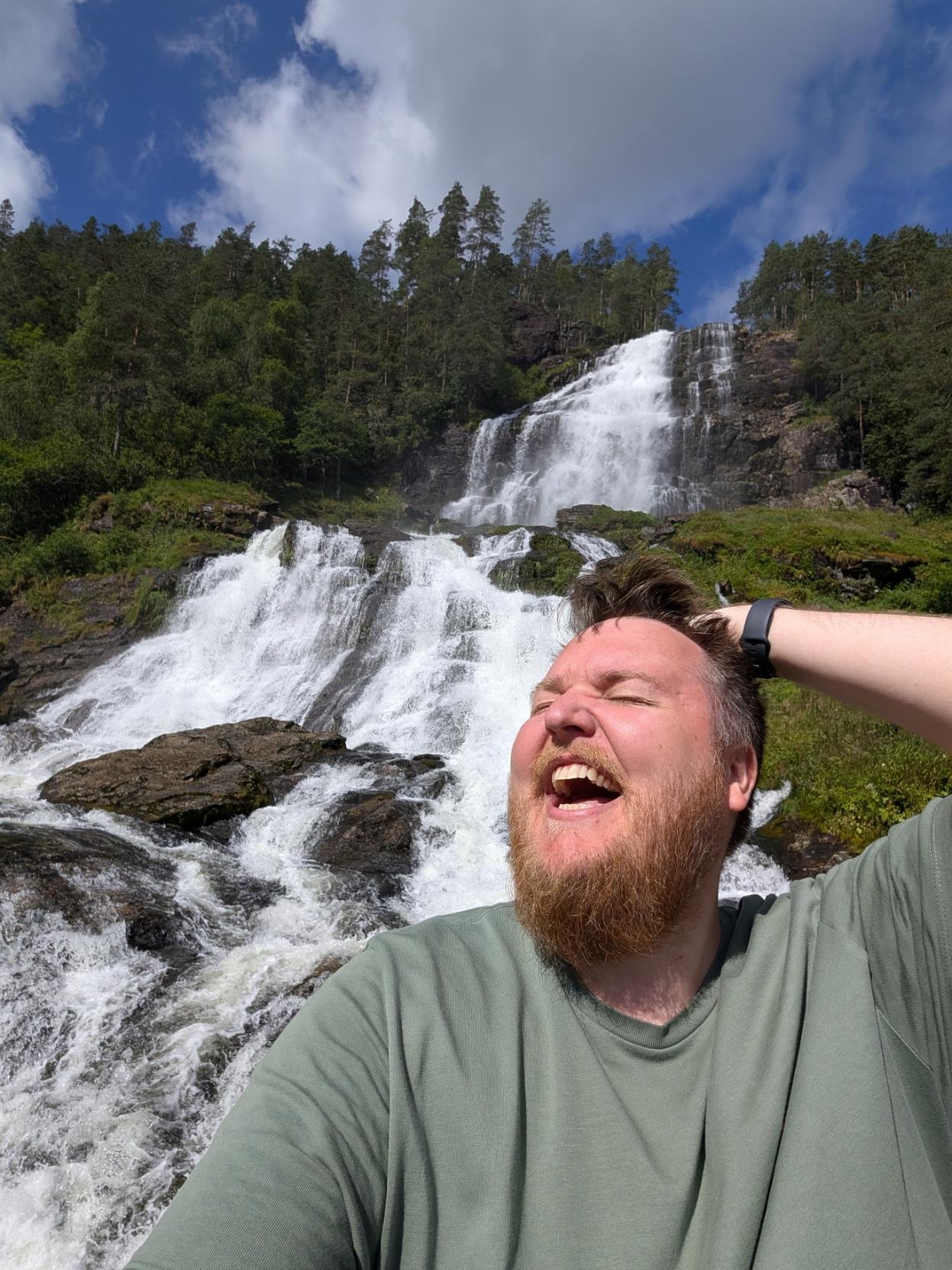Selfie framfor Svandalsfossen: Ein mann med grøn t-skjorte, rødleg skjegg og solbrunt andlet smiler breitt og tek seg til håret med den eine handa. Bak han fossar den mektige Svandalsfossen nedover dei bratte, mose- og skogskledde fjellsidene i fleire trinn. Sola skin, og himmelen over fossen er blå med kvite, lette skyer.