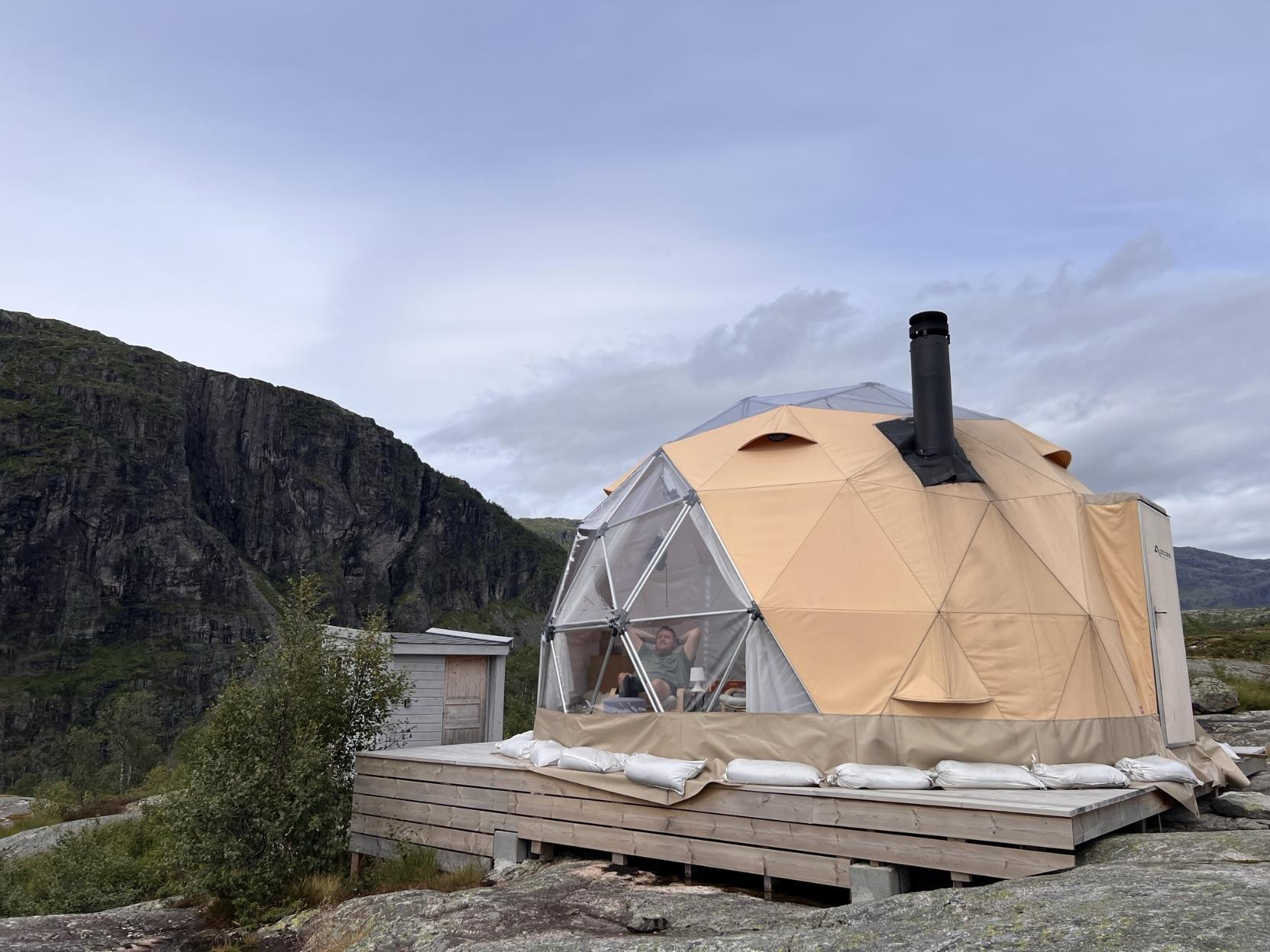 Glamping i fjellet: Ein stor, beigefarga og geodetisk glamping-dome står plassert på ein treplatting på eit svaberg. Den eine sida av domen har eit stort, gjennomsiktig vindauge der ein mann sit inne og slappar av med hendene bak hovudet. Ein svart piperøyr stikk opp frå taket. Bak domen står eit lite trebygg, og i bakgrunnen rager ein bratt, dramatisk fjellvegg under ein overskya himmel.