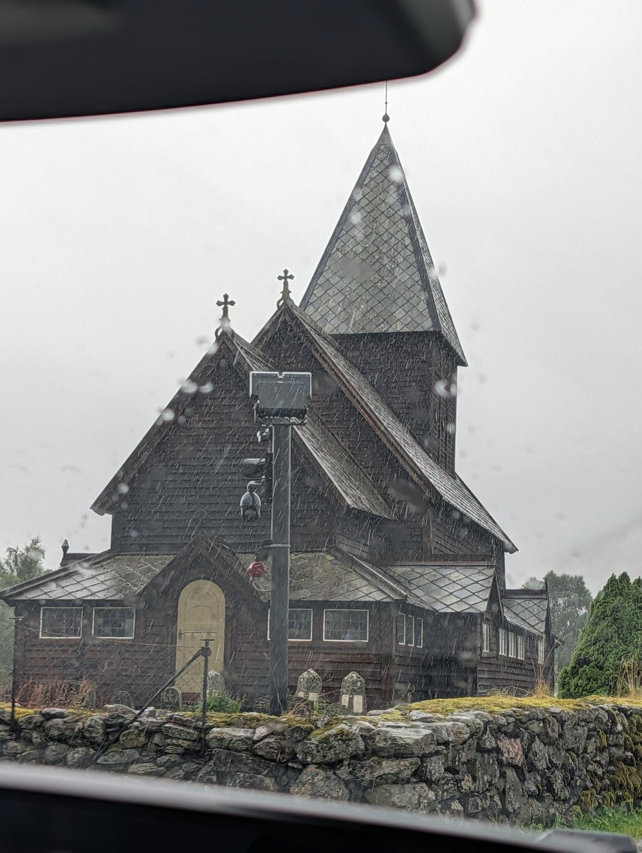 Røldal stavkyrkje i regnvêr: Biletet er teke gjennom ei regndråpete bilrute mot den mørke, historiske trekyrkja. Kyrkja har eit spisst, skifertekt tårn, treshingeltak og krossar på mønene. Framfor kyrkja går ein låg steingard av mosegrodd naturstein, og rett ved inngangen står ein stong med kamera og utstyr. Himmelen i bakgrunnen er grå og overskya.