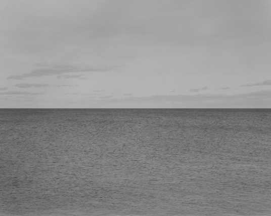 The horizon over a large lake and it is, indeed in the middle. The lake has no large waves or swells but is not smooth and darker areas show where zephyrs of breeze are travelling. Some pale clouds line the horizon but become sparse near us.
