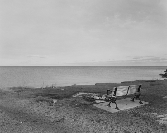 A wooden park bench sit on a concrete pad surrounded by the dry grass of late winter. There are some small patches of snow that has melted to ice. In front of the bench is a makeshift fireplace of gathered stones and it looks out at the unbroken horizon of Lake Ontario.