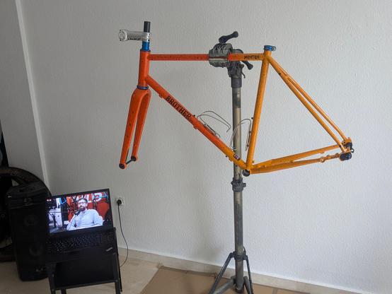Brother Cycles Mehteh gravel bike frame being build. Only the blie anodized headset, seatpost collar, stem and stem spacers have been mounted yet. To the side, a laptop is showing Cade medias "Wild ones podcast" on youtube.