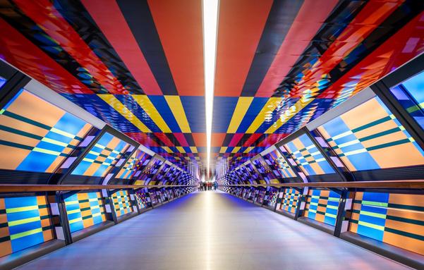 A vibrant, futuristic pedestrian tunnel in Canary Wharf, featuring colorful geometric patterns in red, yellow, blue, and black on the ceiling and angled wall panels. The perspective view leads to a distant group of people at the end of the well-lit corridor, creating a sense of depth and symmetry.