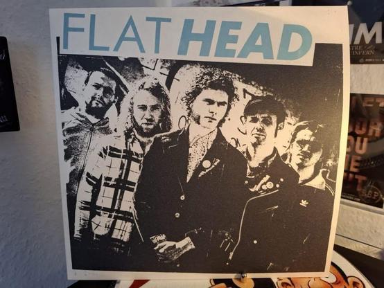 Eine Gruppe von fünf Männern steht in einem stilisierten Schwarz-Weiß-Bild zusammen. Der Text "FLAT HEAD" ist in hellblauer Schrift am oberen Rand zu sehen. Die Männer scheinen unterschiedliche Frisuren, Kleidung und Ausdrücke zu haben, was einen lässigen Eindruck vermittelt.