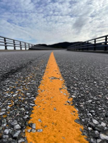 This image features a close-up view of a road surface with a focus on a vibrant yellow road marking that divides the lanes. The perspective is low and close to the ground, making the yellow line lead the viewer's eye towards the horizon. The road is surrounded by guardrails on either side, and the background includes a slightly hazy blue sky with patchy clouds. The texture of the road is prominently displayed, highlighting the rough and speckled asphalt. Hills can be gently seen in the distance, enhancing the depth of the landscape. The overall composition gives a sense of direction and open road journey.