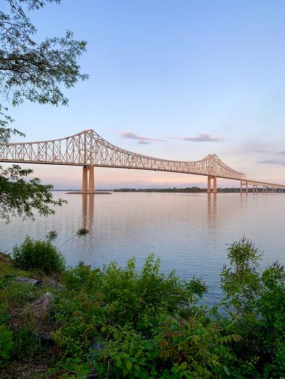 A long cantilever bridge spans a large river in the sunny evening, right before sunset. The far bank is lined with trees, and the near bank has some bushes. The bridge glows in soft orange due to the setting sun. The background is a blue sky with light orange hues close to the horizon.