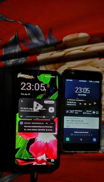 The photograph composed in warm light portrays two smartphones placed on a red fabric with a floral pattern. The phone on the left has a lock screen displaying the time "23:05" and the date "Tue, Apr 29." The background features a vibrant image of a red flower with green leaves. The phone is charging, with a battery level of 82%. A music player widget is visible, showing the song "13_18_21_aug_2021_19Lyntr" by "RadioAzureus (Musicolet)" with a play button and a progress bar. A notification from Mastodon is also visible, mentioning the user by Luca Sironi.

The phone on the right shows a similar time "23:05" and date "Tue, Apr 29." The background is dark blue, and the music player widget displays the same song and artist. The weather widget shows "26°C MonPlaisir" with "Clear" conditions. The battery level is also 82%, and the phone is connected to "GSM provider." Both phones are displaying the same time, indicating they are synchronized. The Android on the right is in slight Bokeh due to the shallow DOF because of the wide aparture 

 Ovis2-8B

🌱 Energy used: 0.266 Wh