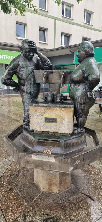 A humorous bronze fountain sculpture featuring two life-sized figures, a man and a woman, standing on either side of a large water basin with mugs beneath small spouts. The man appears frustrated, holding his head in one hand, while the woman looks sternly at him. A sign on the fountain reads “Kein Trinkwasser” (No drinking water). The scene is set on a wet pavement in front of a building with green signage.