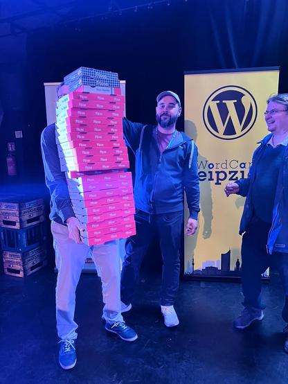 Stapel mit Pizzen, getragen von einem Mann, bewundert von 2 weiteren vor dem Flyer vom WordCamp Leipzig