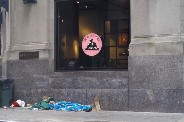 A window with a "Joe & The Juice" sign depicting a man in suit with his head leaning on his right fist, with his elbow on the window. Under the window is an unhoused woman, sleeping with blankets, with her right hand raised to her face.