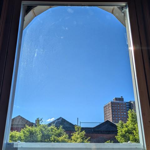 Looking through an arched window two and a half hours after sunrise the sky is bright blue, with one small puffy white cloud just above the horizon. Pointed roofs of Harlem brownstones are silhouetted across the street, and a taller apartment building can be seen in the distance. The green tops of two trees are on the bottom and right.