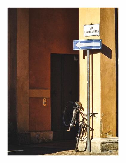 This photo is of a quiet street corner in the afternoon with warm sunlight casting long shadows. There bicycle is parked badly with its back wheel off the ground and locked to a pole. At the top of the pole there is a one-way street sign pointing left. Above this again there is a street name sign that reads "Via Santa Caterina." The bicycle, signs and pole are on the right hand side of the photo in the sunlight, the rest of the photo is in the shade.