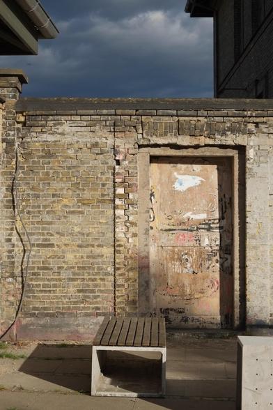 Photograph of a distressed brick wall in a former wholesale harbour district. There is a closed wooden door in the wall that is covered with layers of removed posters and graffiti. A newer rectangular city bench of concrete with wooden seating slats is in front of the door. Tufts of grass stick out between the concrete slabs on the ground. The sun is low and casting strong angular shadows. Behind the wall the sky is dark and cloudy, indicating a coming storm. Kødbyen, Vesterbro, Copenhagen, Denmark, 30 June 2018. Photo Cindy Kohtala