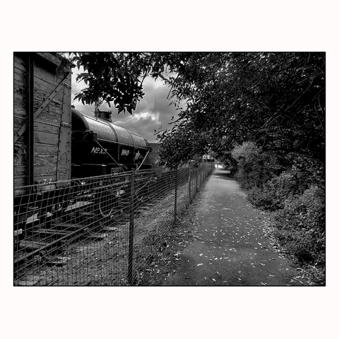 A railway line and footpath are separated by a metal wire fence, there are antique boxcars and containers held in storage whilst both lead toward the city and wapping wharf.