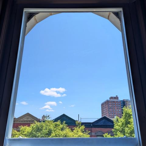 Looking through an arched window at midday the sky is bright blue with a few small puffy white clouds. Pointed roofs of Harlem brownstones with red brickwork are across the street, and a taller apartment building can be seen in the distance. The green tops of two trees are on the bottom and right.
