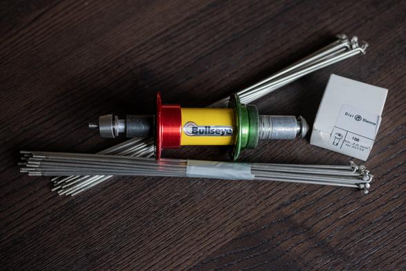 Photo of a bicycle rear hub, two bundles of silver spokes, and a white box on a brown wooden table. The hub is tricolour with a red flange, a green flange, and a yellow middle body. A label on the boy says 'Biel Bienne 100 für 2mm Nr. 65133'.