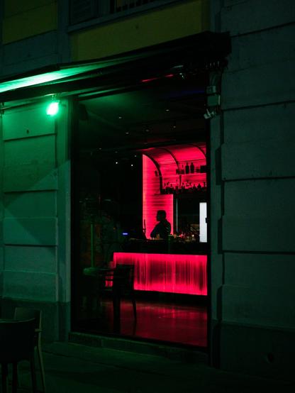 The silhouette of a man is partly visibile inside a bar whose furniture is lit with red neon light. The outside of the bar has a green light casting a greenish tone to the scene.