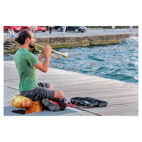 a person playing a trumpet while sitting near a wooden boardwalk by the water. The individual is dressed casually in a green t-shirt and shorts. An open instrument case lies on the boardwalk in front of them, suggesting they might be busking or simply enjoying playing music outdoors. The background shows a body of water with waves gently crashing against a stone barrier, and there are people walking and cars parked in the distance. The scene suggests a relaxed, coastal environment.