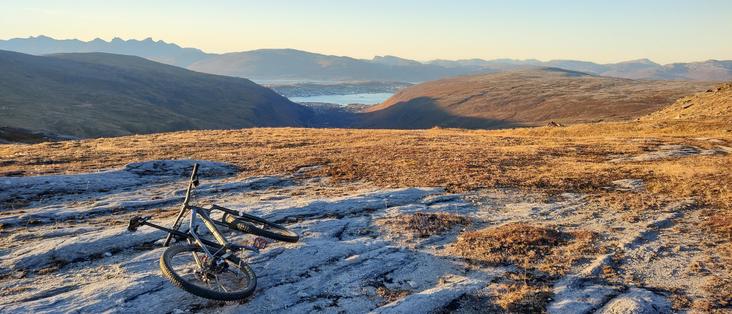 A bike sits atop a rock slab overlooking a valley and fjord. Moments before a wild descent of the Tromsdalstind sommerruta 🙌