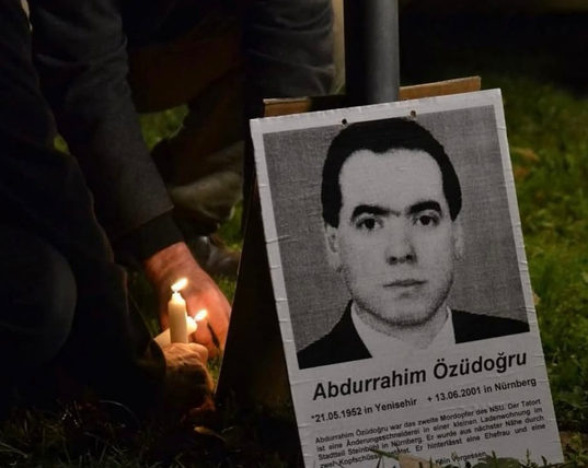 Ein Plakat mit Portrait von Abdurrahim Özüdoğru lehnt an einem Pfosten. Unter dem Bild stehen einige Informationen zu ihm. Links daneben sieht man Hände, die Kerzen anzünden.