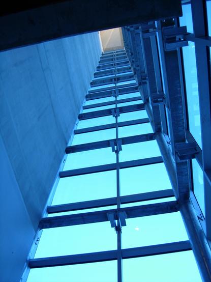 Color photo looking up through an open shaft that is defined by a smooth concrete wall (left) and two walls of metal framed blue glass (center and right)