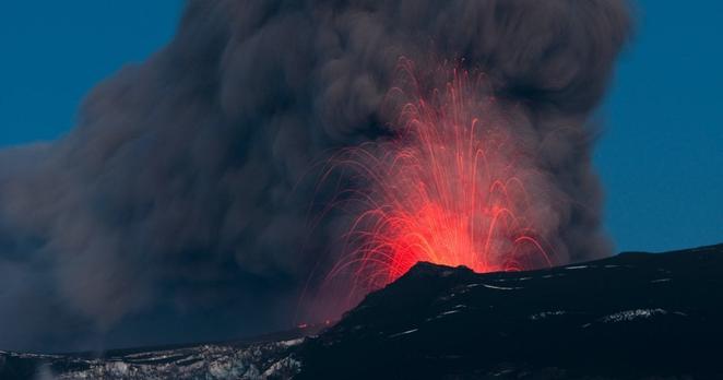 Ein schwarzer oder schwarz berußter Berg, aus dessen Spitze glühend rote Fontänen in die Höhe schießen, durch die Bewegungsunschärfe in weiten Bögen. Links darunter ist ein sehr schmutzig grauer Gletscher zu sehen. Der Himmel ist abgesehen von dunkelgrauem Rauch blau.