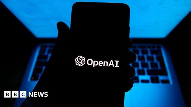 A person holding a phone with OpenAI software in a dark room with a computer behind their hand