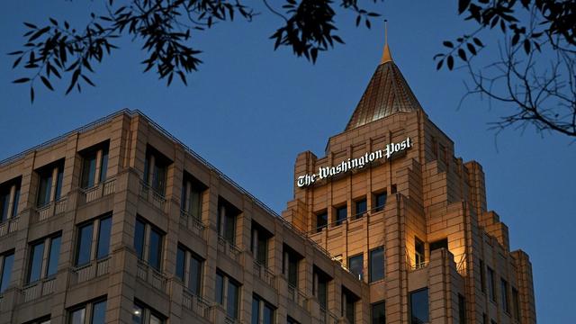 Exterior of The Washington Post building at dusk, with lights on inside some windows and tree branches framing the top against a clear blue sky.