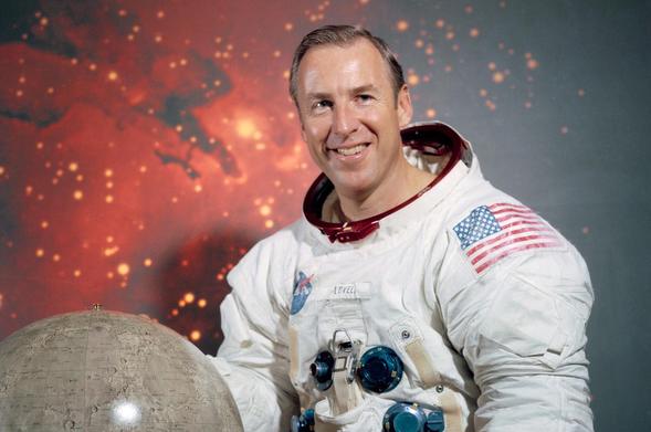 NASA astronaut Jim Lovell smiles in a white astronaut suit with a shiny, gray globe and a picture of stars in deep space behind him.