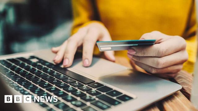 A person sits at a laptop computer. We can see a close-up of their hands. They are holding a credit card, and it appears that they are typing its numbers into the computer.