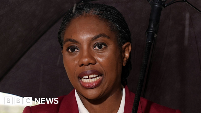 Headshot of Kemi Badenoch, who has her black hair tied back. She is holding a black umbrella and wearing a burgundy jacket with matching lipstick and a whote collared shirt, without jewellery.