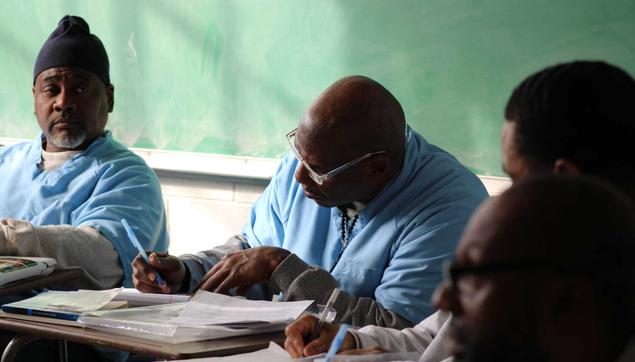 Men in blue shirts sit at desks in front of a green chalkboard inside a classroom within Sheridan Correctional Center.