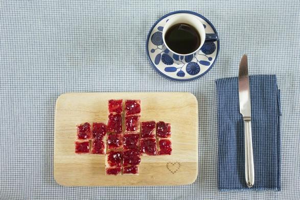 Auf hellblauer Tischdecke: Brettchen mit Marmeladentost in Kreuzform, Tasse Kaffe in Blümchenporzellan, Messer auf karierter Serviette,