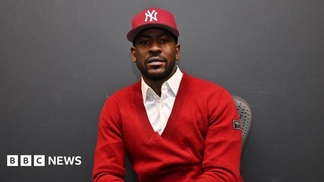 A UK grime artist wearing a red jumper with a white shirt underneath. He is wearing a red cap and looking directly at the camera.