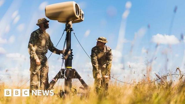 Two men in military khaki use a bit of kit that is on a stand