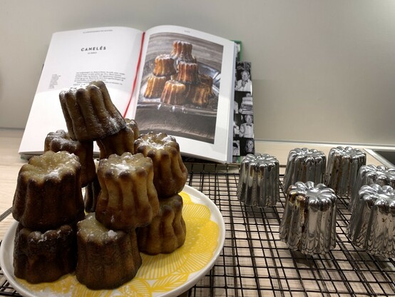 Kjøkkenbenk med bakerist. Stabel med 12 småkaker (caneles de Bordeaux) på et gult fat, seks aluminiumsformer, kokebok i bakgrunnen