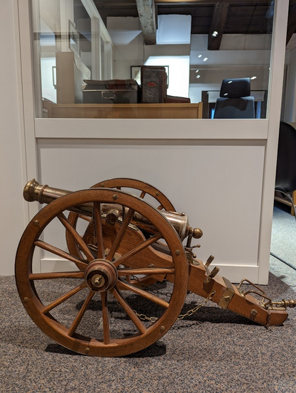 En liten, messingfarget kanon på en nydelig tre-lavett med to store hjul står foran et kontor.