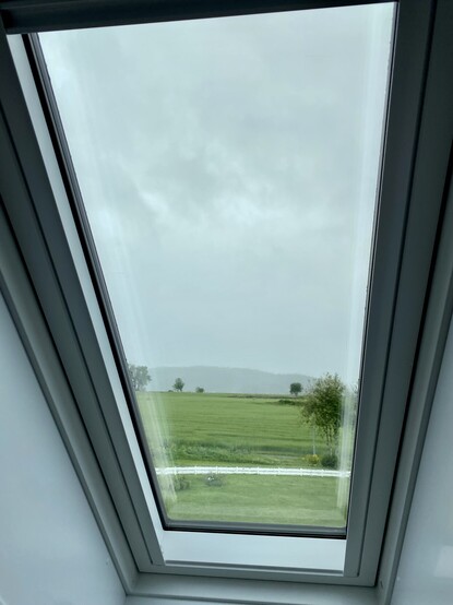 View from a roof window. A lush green field and scattered trees. The sky is heavy and grey.