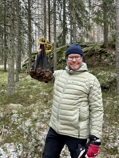 Mann med lue og dunjakke i skogsomgivelser viser et soppnett med noen brune sopp i.