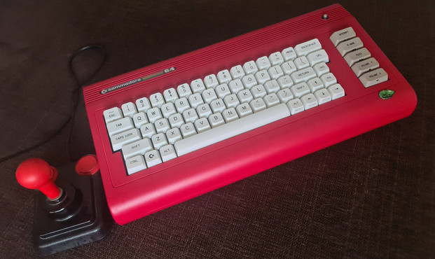 On a sofa, a shiny, red Commodore 64X computer case sporting a white Cherry MX mechanical keyboard is looking seductively at the camera. Next to it, a 40 year old Competition Pro 5000 joystick, begging for a date with Daley Thompson's Decathon.
