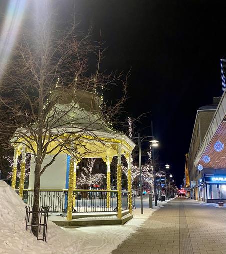 En gul paviljong, et tre og en stor haug med snø opplyst av gatelys i hovedgata i byen. Hovedgata fortsetter innover i bildet og er kranset med butikker og lyspyntede trær.