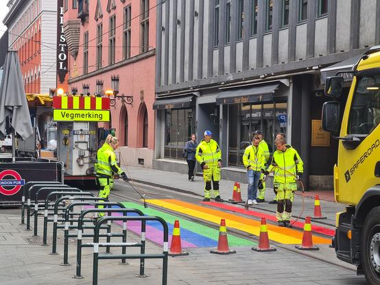 Arbeidere varmer opp vegmerking i regnbuens farger for å feste den til gata. En arbeidsbil merket "Vegmerking" står bak.