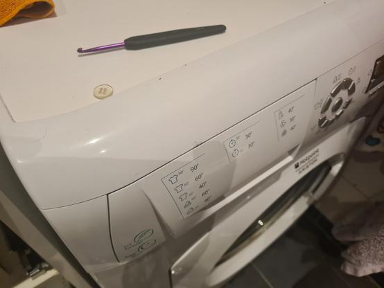 Utsnitt av vaskemaskin. På toppen ligger en knapp og ei heklenål. Merket på maskinen vises over døra, Hotpoint Ariston.