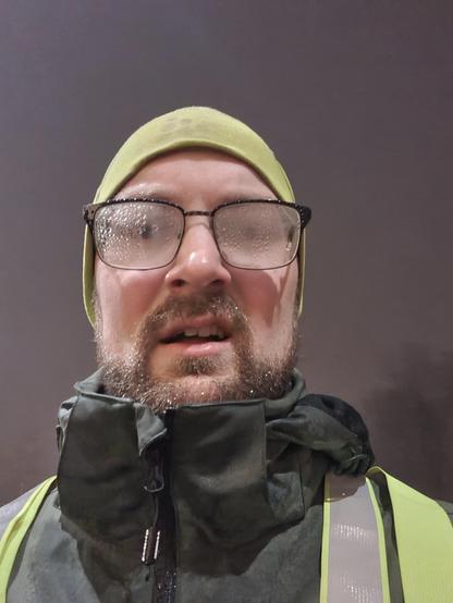 Ansiktet mitt, regnvått, dugg og regndråper på brillene, konsentrert ansiktsuttrykk. Gul lue, regnjakke og refleksene.