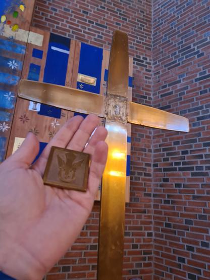 Prosesjonskors i messing. I krysset mellom korsbjelkene er det er stygt felt. Hånda mi holder opp det kvadratiske feltet med et due-relieff som har sittet der, men som har falt av. I bakgrunnen ses litt av altertavla og murveggen.