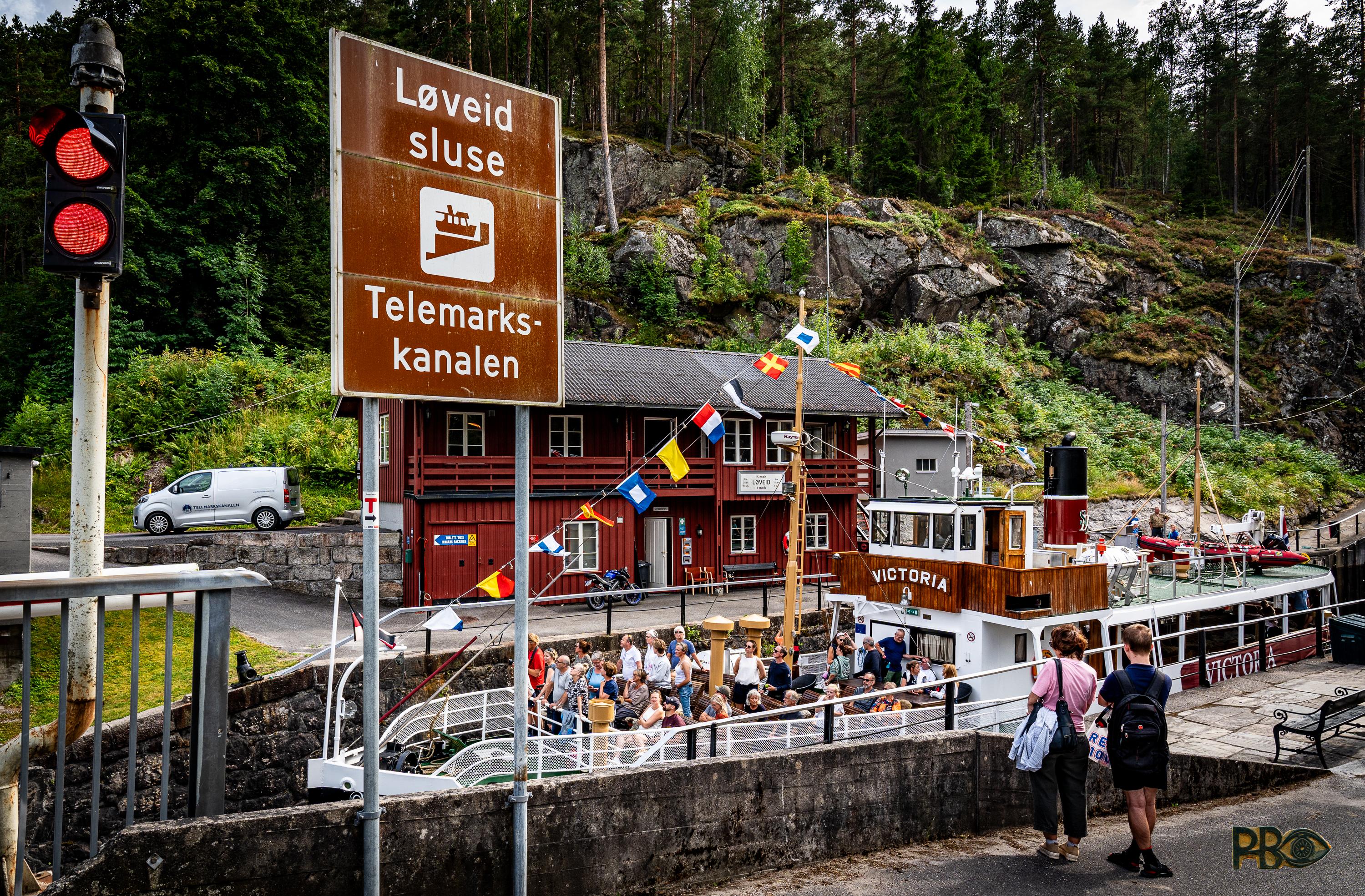 Victoria med sine mange vimpler er på vei ut fra slusene , nå er den på nivå med Meierelva ved Skotfoss. På fordekket sitter mange reisende og er vel egentlig ganske trøtte nå etter den lange turen over Norsjø. I forgrunnen høyt til venstre står et stort skilt som forteller: Løveid sluse, Telemarkskanalen. Høyt helt til venstre knallrødt trafikklys som forteller at brua over kanalen står oppe.
