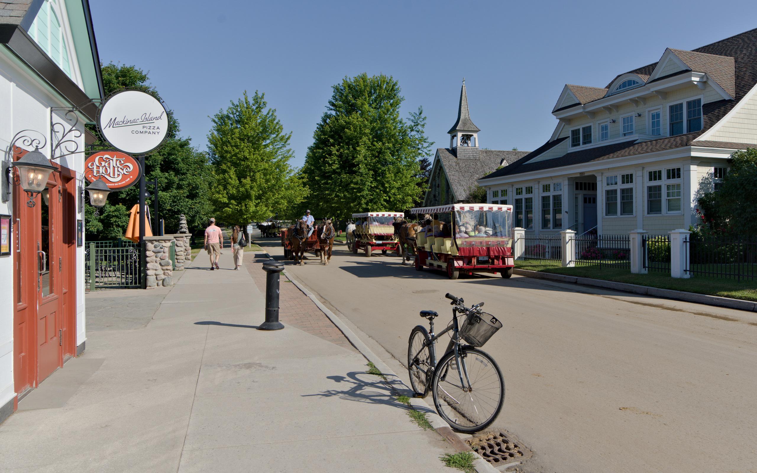Street scene from Mackinac Island: A bicycle is parked in the forground in front of the Mackinac Island pizza company store. Two people are walking away from the camera on the sidewalk. A two-horse carriage is approaching and two covered horse-drawn carriages are going down the opposite side of the boulevard. The boulevard is lined with green trees.