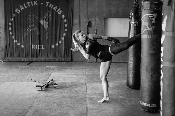 Junge Frau in einer Kickbox-Halle beim Training. Sie hat am rechten Bein einen Oberschenkelgips und kickt mit dem Gips hoch gegen einen Sandsack. Auf dem Boden liegen ihre Krücken.